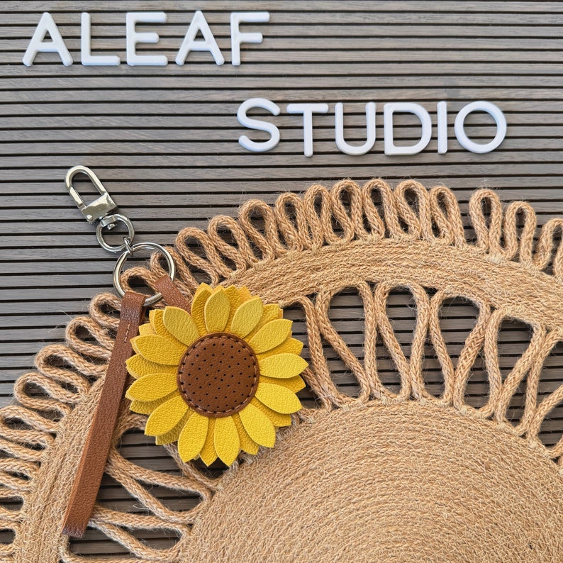 May include: A yellow sunflower keychain with a brown center and a brown leather strap attached to a silver key ring. The keychain is on a brown woven mat with the words "ALEAF STUDIO" in white letters on a gray background.