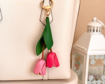 Puede incluir: Un bolso tote de cuero blanco con una cadena dorada y un llavero de cuero rosa y verde en forma de tulipán.
