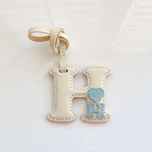 Puede incluir: Una etiqueta de cuero color crema con forma de letra H y una correa de lazo. La letra presenta un peque&ntilde;o coraz&oacute;n azul claro y una letra H m&aacute;s peque&ntilde;a en azul claro cosidas en la parte inferior derecha. Los bordes tienen costuras color crema.