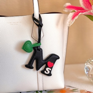 Puede incluir: Un bolso tote de cuero granulado blanco con una correa de cuero negra colgando del asa. La correa presenta un dije de letra M negra, un dije de coraz&oacute;n verde peque&ntilde;o, un aplique de coraz&oacute;n rojo y un aplique de letra S blanca sobre la M negra.