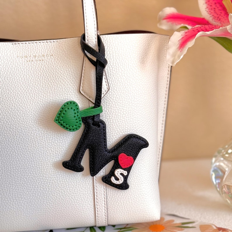 Puede incluir: Un bolso tote de cuero granulado blanco con una correa de cuero negra colgando del asa. La correa presenta un dije de letra M negra, un dije de coraz&oacute;n verde peque&ntilde;o, un aplique de coraz&oacute;n rojo y un aplique de letra S blanca sobre la M negra.