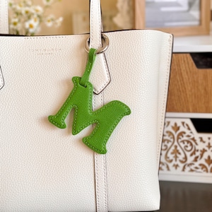 May include: A white leather tote bag with a green leather letter "M" charm hanging from the handle. The charm is attached with a silver ring. The bag has a simple, elegant design.