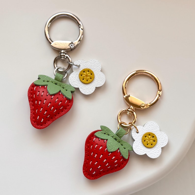 Puede incluir: Dos llaveros con un dije de fresa roja con hojas verdes y un dije de flor blanca con centro amarillo. Cada fresa tiene detalles de costura en blanco. Los llaveros est&aacute;n sujetos a anillas de metal en tonos plateado y dorado.