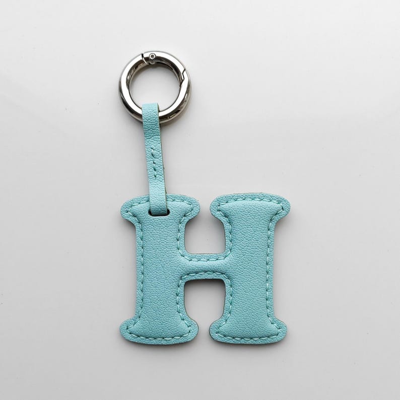 May include: A light blue leather keychain in the shape of the letter "H". The keychain has a silver-colored metal clasp and a matching leather strap. The edges of the letter are stitched with a contrasting light-colored thread.