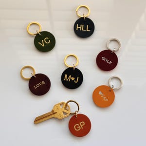 Pode incluir: Uma coleção de chaveiros personalizados em várias cores, incluindo verde, preto, bordô e laranja. Cada chaveiro apresenta um anel dourado ou prateado e é gravado com iniciais ou palavras como "LOVE" e "GOLF". Uma chave dourada também está presente.