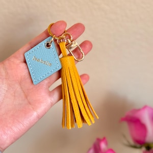 May include: A light blue leather keychain with the name "Isabelle" embossed on it. The keychain has a gold metal ring and a yellow tassel.