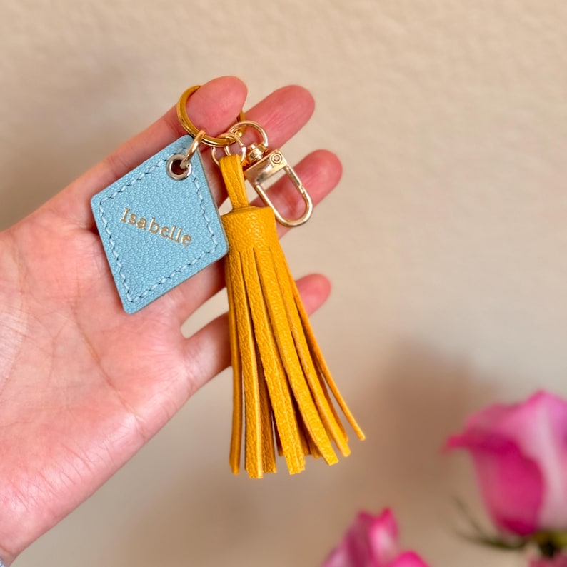May include: A light blue leather keychain with the name "Isabelle" embossed on it. The keychain has a gold metal ring and a yellow tassel.