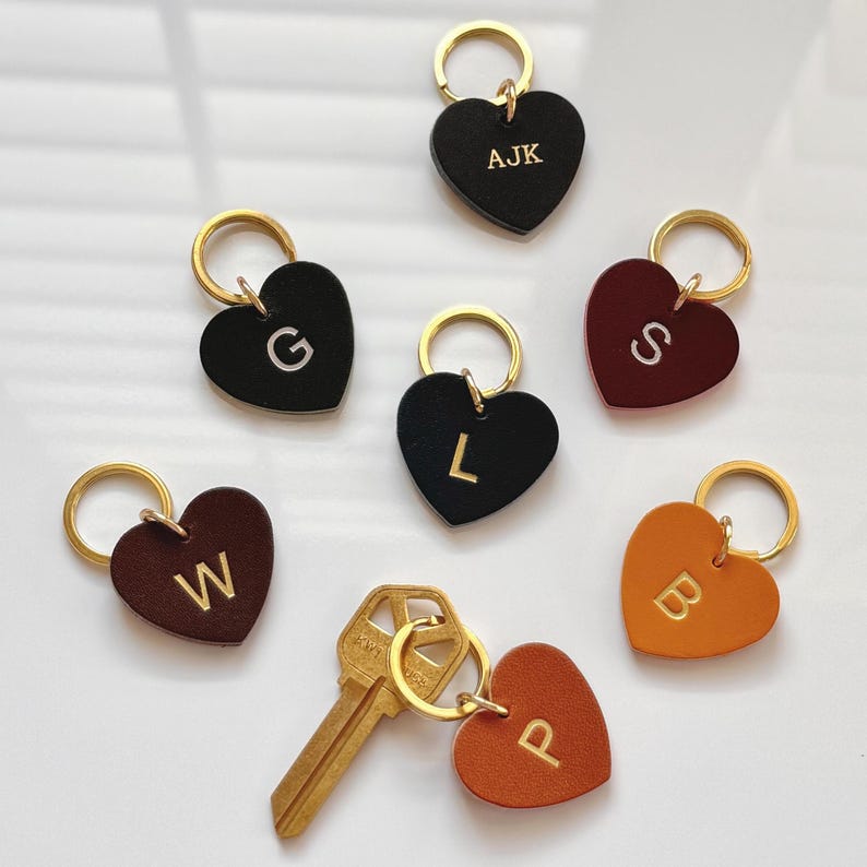 May include: Heart-shaped keychains in various colors, including black, brown, and orange, each with a gold-colored keyring. The keychains feature gold-colored letters: AJK, G, L, S, W, B, and P. A gold key is also present.