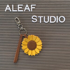 May include: A brown leather keychain with a silver key ring and a yellow felt sunflower charm. The keychain is hanging on a gray pegboard with the words "ALEAF STUDIO" in white letters.