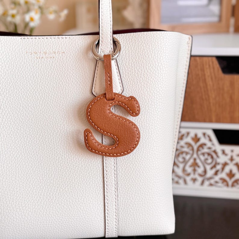 May include: A white leather tote bag with a brown leather letter "S" charm hanging from the handle. The charm is attached to a silver ring. The bag has a minimalist design with a subtle embossed logo.