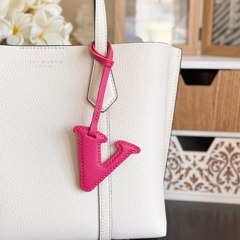 May include: A white leather tote bag with a pink leather letter "V" charm hanging from the handle. The bag has a textured surface and a simple design. The charm is a bright, eye-catching accent.