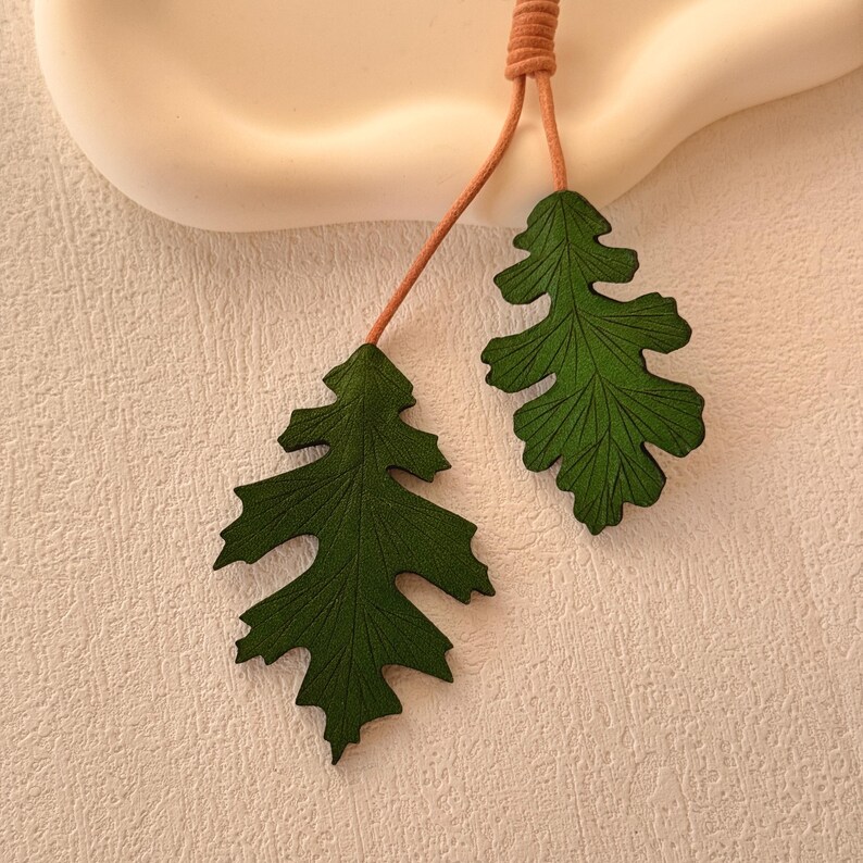 May include: Two green leather leaves with detailed veins hanging from a brown cord. The leaves are a dark green color and have a textured surface.