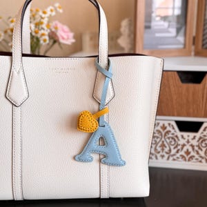 Puede incluir: Un bolso tote de cuero granulado blanco con el logotipo de Tory Burch grabado en la parte delantera. Una correa de cuero azul claro cuelga del asa, con un dije de la letra A en azul claro y un peque&ntilde;o coraz&oacute;n amarillo con costuras visibles.
