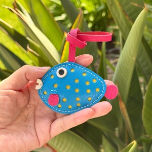 Puede incluir: Un llavero de cuero azul en forma de pez globo con lunares amarillos, una lengua rosa y un ojo blanco. El llavero tiene una correa de cuero rosa.