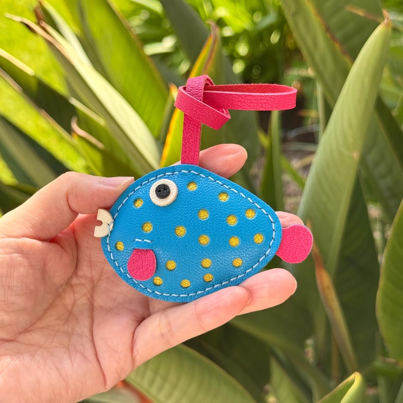 Puede incluir: Un llavero de cuero azul en forma de pez globo con lunares amarillos, una lengua rosa y un ojo blanco. El llavero tiene una correa de cuero rosa.