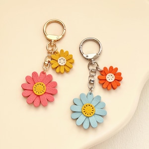 Op de afbeelding: Twee sleutelhangers in de vorm van madeliefjes. Eén sleutelhanger heeft een roze bloem met een gele kern en een kleinere gele bloem. De andere sleutelhanger heeft een blauwe bloem met een gele kern en een kleinere oranje bloem. Beide hebben metalen sleutelringen.