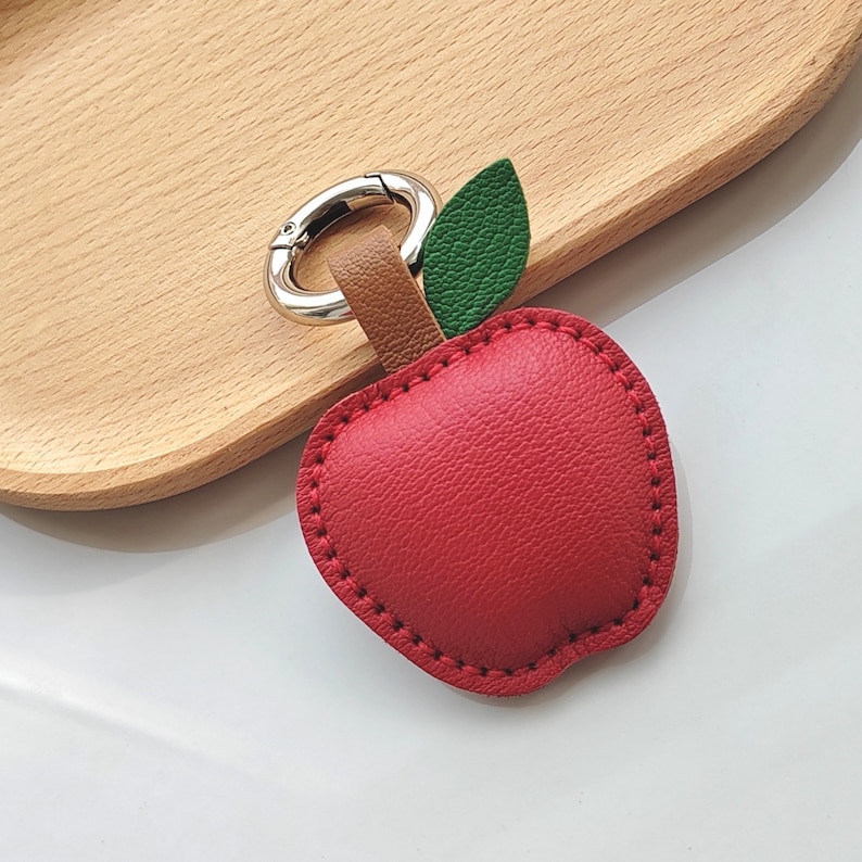 Puede incluir: Un llavero de cuero rojo en forma de manzana con una hoja verde y un anillo de metal plateado.