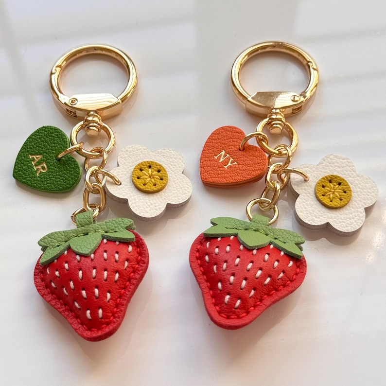 Puede incluir: Dos llaveros de metal dorado con un dije de fresa roja con hojas verdes, una flor blanca con centro amarillo y un dije en forma de coraz&oacute;n. Un coraz&oacute;n es verde con las letras AR y el otro es naranja con las letras NY grabadas.