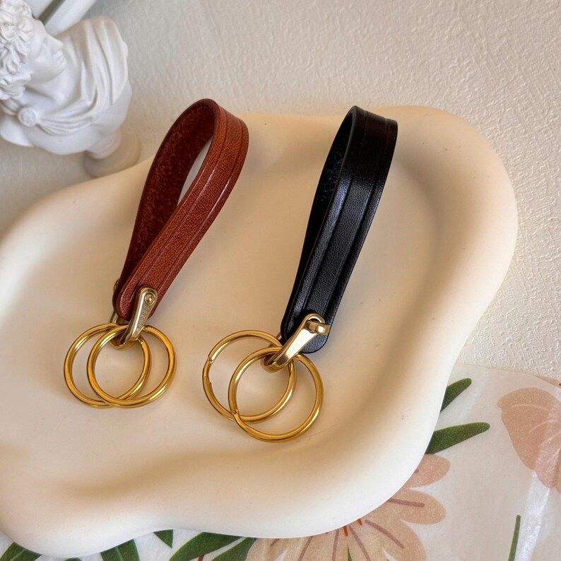 May include: Two leather keychains with gold double rings. One keychain is brown leather and the other is black leather.
