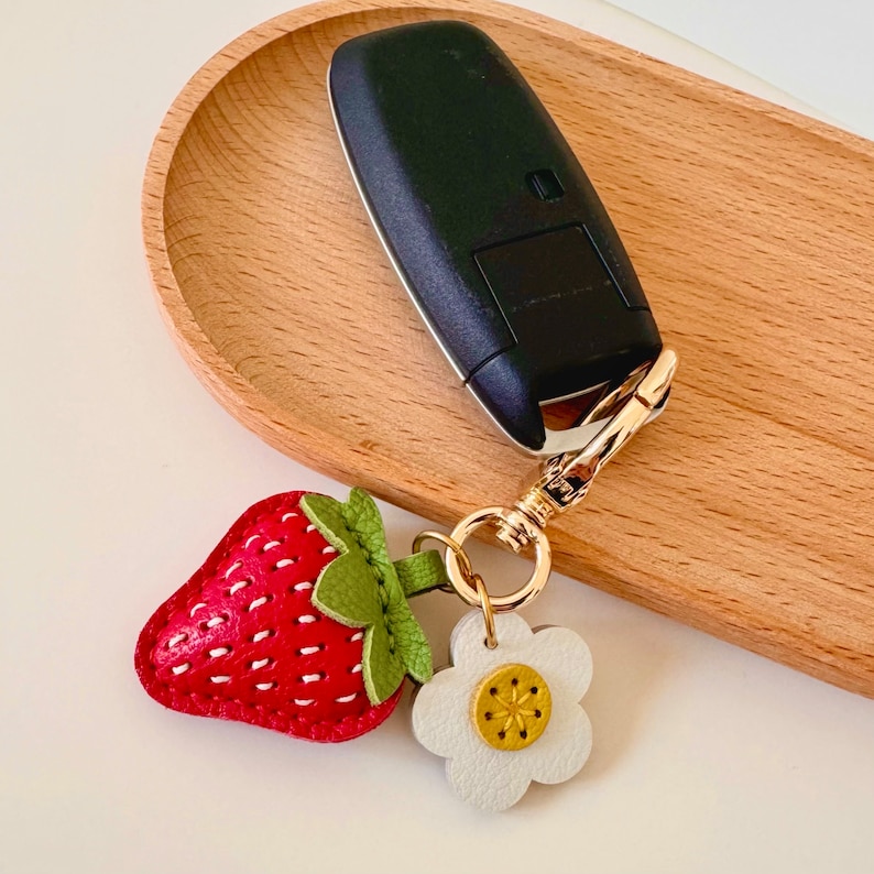 May include: A red leather strawberry keychain with a white leather flower charm and a gold metal key ring. The keychain is attached to a black car key.