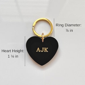 May include: Black heart-shaped keychain with gold-colored ring. The heart measures 1 ⅛ inches in height and features the initials "AJK" in gold. The ring diameter is ⅞ inch. The keychain is a personalized accessory.