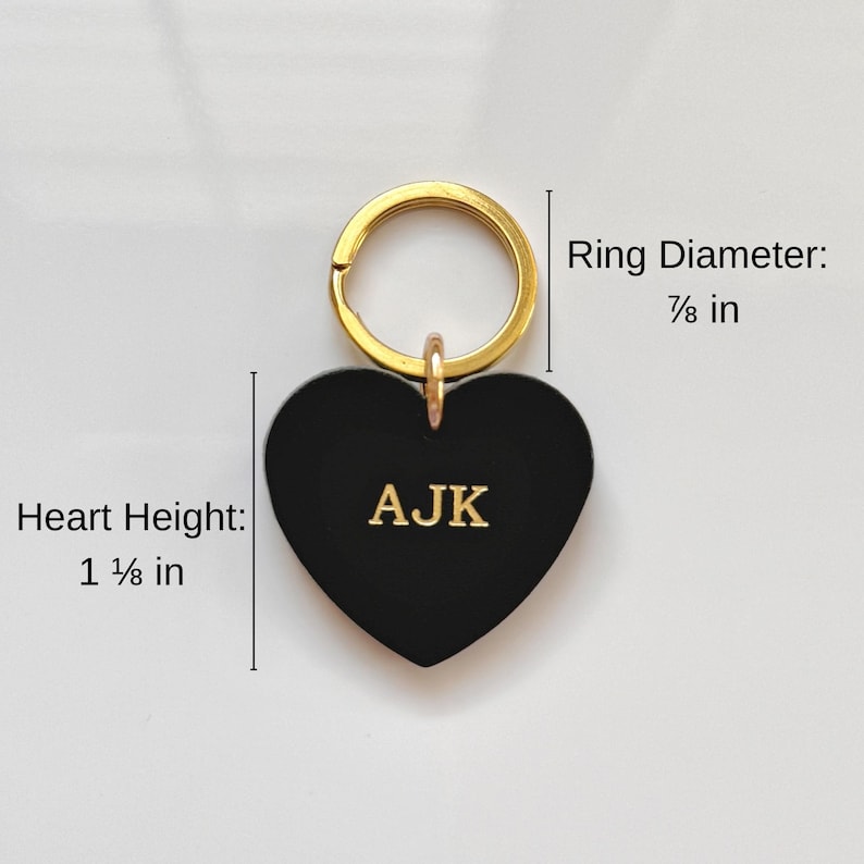 May include: Black heart-shaped keychain with gold-colored ring. The heart measures 1 ⅛ inches in height and features the initials "AJK" in gold. The ring diameter is ⅞ inch. The keychain is a personalized accessory.