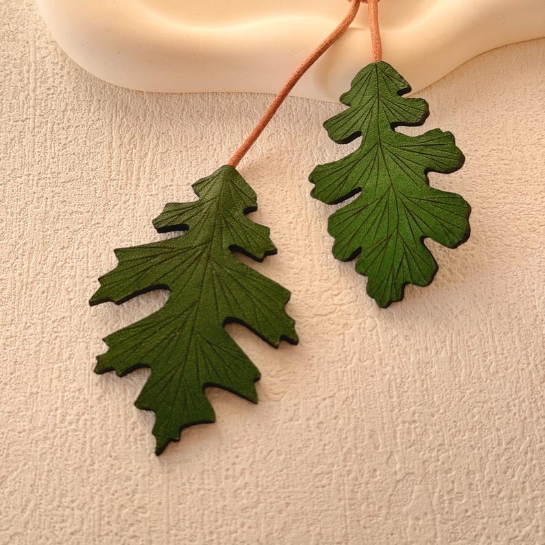 May include: Two green leather leaf earrings with detailed veins. The leaves are attached to brown cords.