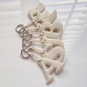 May include: Off-white leather keychains spelling out the word "ABBAS". Each letter is individually cut and stitched, with a silver-colored metal ring attached for keys. The keychains are arranged vertically.