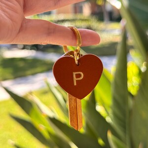 May include: A gold-colored key ring holding a brown leather heart-shaped tag with the letter "P" in gold. A gold-colored key is attached to the ring. The background is blurred with green foliage.