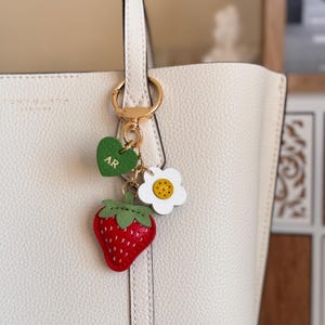 Puede incluir: Un llavero de metal dorado sujeto a un bolso de mano texturizado de color crema. El llavero presenta un dije de fresa roja con hojas verdes, una flor blanca con centro amarillo y un coraz&oacute;n verde grabado con las letras AR.
