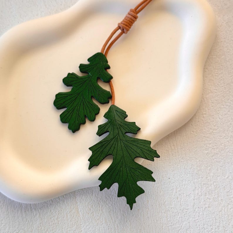 May include: A green leather necklace with two leaf pendants. The leaves are detailed with veins and have a textured surface.