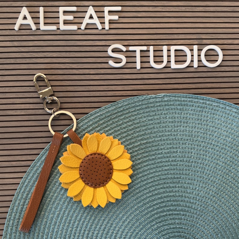 May include: A brown leather keychain with a sunflower charm. The sunflower is yellow with a brown center and has a textured leather look. The keychain has a silver metal ring and a silver metal clasp.