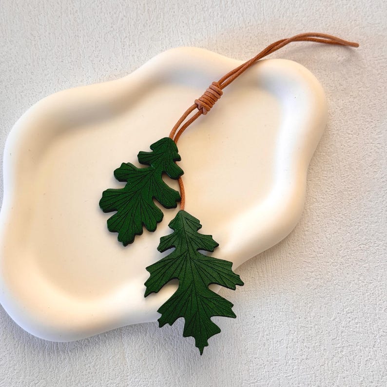 May include: Two green leather leaves attached to a brown leather cord. The leaves are detailed with a textured pattern.