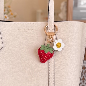 Puede incluir: Un bolso tote de cuero color crema con el texto Tory Burch New York grabado en la parte delantera. Enganchado al asa hay un llavero de metal dorado con un dije de fresa roja con hojas verdes y una flor blanca con el centro amarillo.