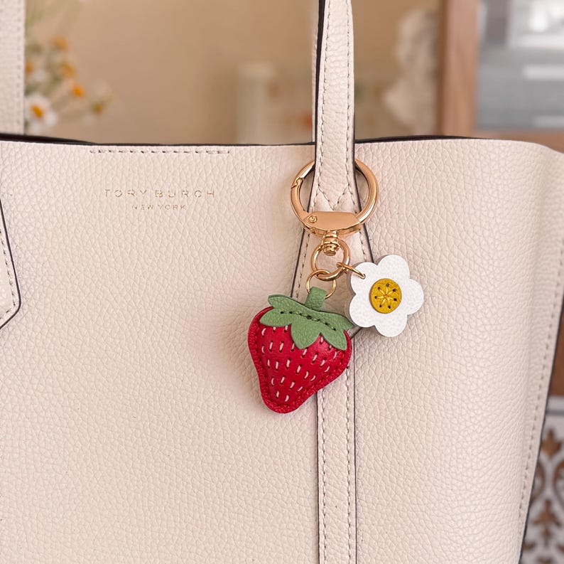 Puede incluir: Un bolso tote de cuero color crema con el texto Tory Burch New York grabado en la parte delantera. Enganchado al asa hay un llavero de metal dorado con un dije de fresa roja con hojas verdes y una flor blanca con el centro amarillo.