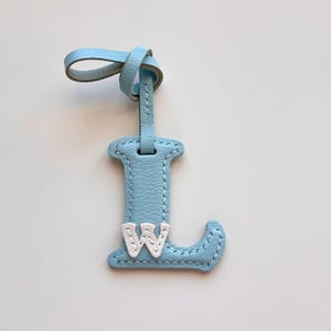 Peut inclure: Un porte-étiquette de bagage en cuir bleu clair en forme de la lettre "L", avec un "W" blanc cousu en bas. Il possède une boucle bleue assortie pour l'attache. L'étiquette est en cuir et présente une bordure cousue, idéale pour les voyages.