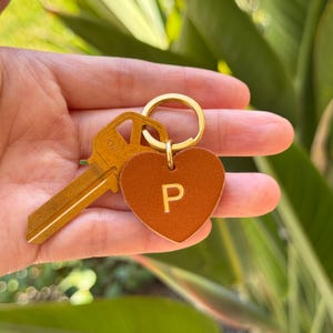 May include: A gold-colored key and a brown leather heart-shaped keychain with the letter "P" in gold. The keychain is attached to a gold ring.