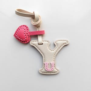Puede incluir: Un llavero de cuero color crema con forma de letra Y, que presenta un peque&ntilde;o dije de coraz&oacute;n rosa unido por una correa y una letra U de color rosa claro cosida en el centro de la Y. El llavero incluye un lazo para su sujeci&oacute;n.