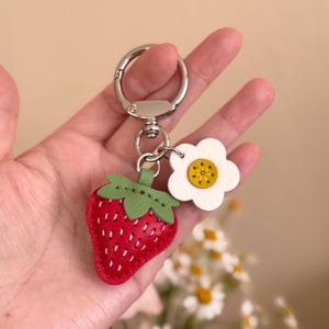 Puede incluir: Un llavero que presenta un dije de fresa roja con hojas verdes y costuras blancas, junto a un peque&ntilde;o dije de flor blanca con el centro amarillo. Los dijes est&aacute;n sujetos a una argolla y un broche giratorio de metal color plateado.