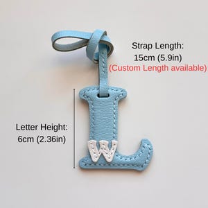 Puede incluir: Una etiqueta de cuero azul claro con forma de letra L y una letra W blanca cosida en la parte frontal. La etiqueta incluye una correa. El texto indica una longitud de correa de 15 cm y una altura de letra de 6 cm, con medidas personalizadas.
