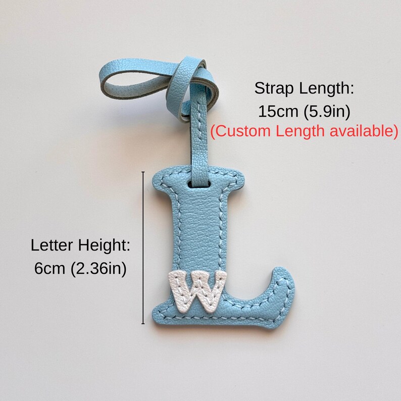 Puede incluir: Una etiqueta de cuero azul claro con forma de letra L y una letra W blanca cosida en la parte frontal. La etiqueta incluye una correa. El texto indica una longitud de correa de 15 cm y una altura de letra de 6 cm, con medidas personalizadas.