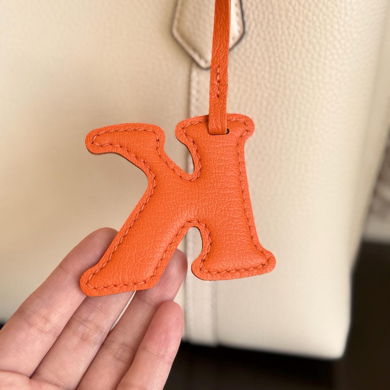 May include: Close-up of an orange leather letter "K" charm with a matching strap, hanging in front of a cream-colored leather bag. The charm has a stitched border and is likely a bag accessory.