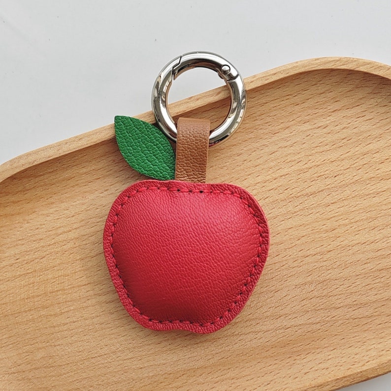 Puede incluir: Un llavero de cuero rojo en forma de manzana con una hoja verde y un anillo de metal plateado.