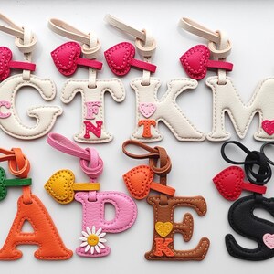 Puede incluir: Una colecci&oacute;n de doce llaveros de cuero con letras del alfabeto en varios colores, incluyendo rosa, blanco, azul, naranja, marr&oacute;n, negro y verde oscuro. Cada letra presenta un peque&ntilde;o dije de coraz&oacute;n o flor y un lazo de cuero para colgar.