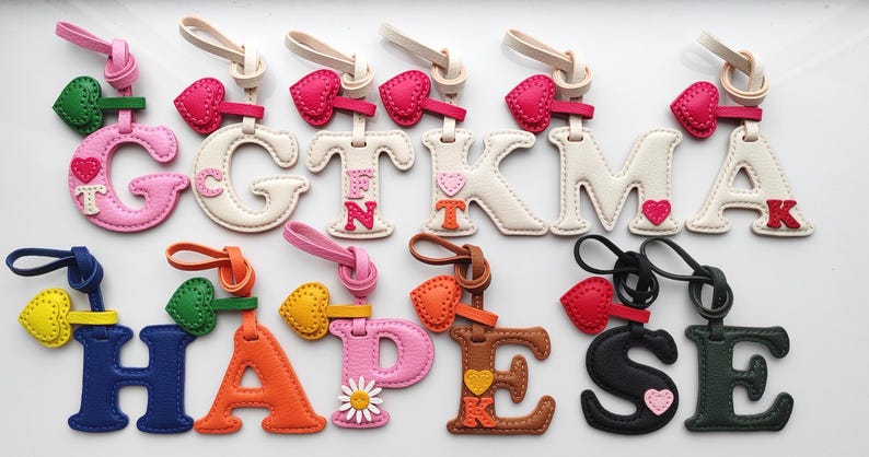 Puede incluir: Una colecci&oacute;n de doce llaveros de cuero con letras del alfabeto en varios colores, incluyendo rosa, blanco, azul, naranja, marr&oacute;n, negro y verde oscuro. Cada letra presenta un peque&ntilde;o dije de coraz&oacute;n o flor y un lazo de cuero para colgar.
