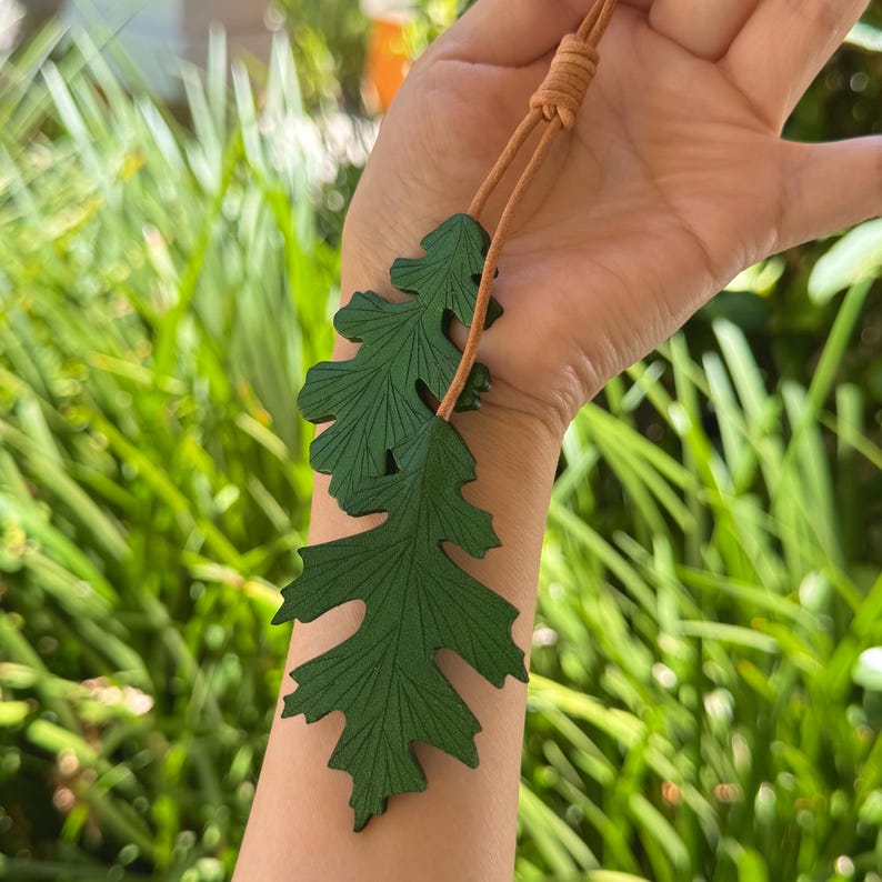 May include: A green leather leaf pendant necklace with a brown leather cord. The pendant is made of two leaves, one on top of the other. The leaves have a detailed vein pattern.
