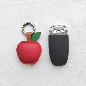 Puede incluir: Un llavero de cuero rojo en forma de manzana con una hoja verde y un anillo plateado. El llavero est&aacute; al lado de un llavero de coche negro.