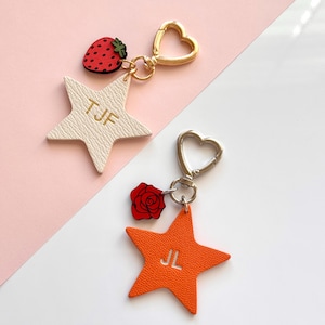 May include: Two star-shaped keychains with gold and silver heart-shaped clasps. One is white with "TJF" and a strawberry charm. The other is orange with "JL" and a red rose charm. Both have textured surfaces.