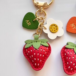 Puede incluir: Un llavero de metal dorado con un dije de fresa roja con costuras blancas y hojas verdes, una flor blanca con centro amarillo y un coraz&oacute;n verde grabado con las letras doradas AR, todo unido a una argolla circular.
