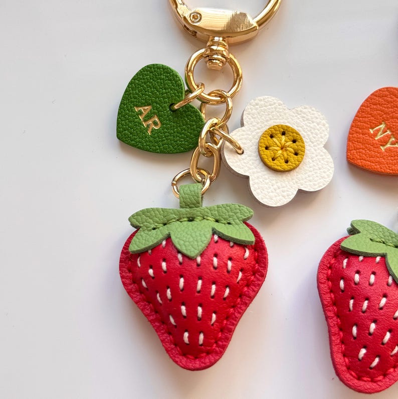 Puede incluir: Un llavero de metal dorado con un dije de fresa roja con costuras blancas y hojas verdes, una flor blanca con centro amarillo y un coraz&oacute;n verde grabado con las letras doradas AR, todo unido a una argolla circular.